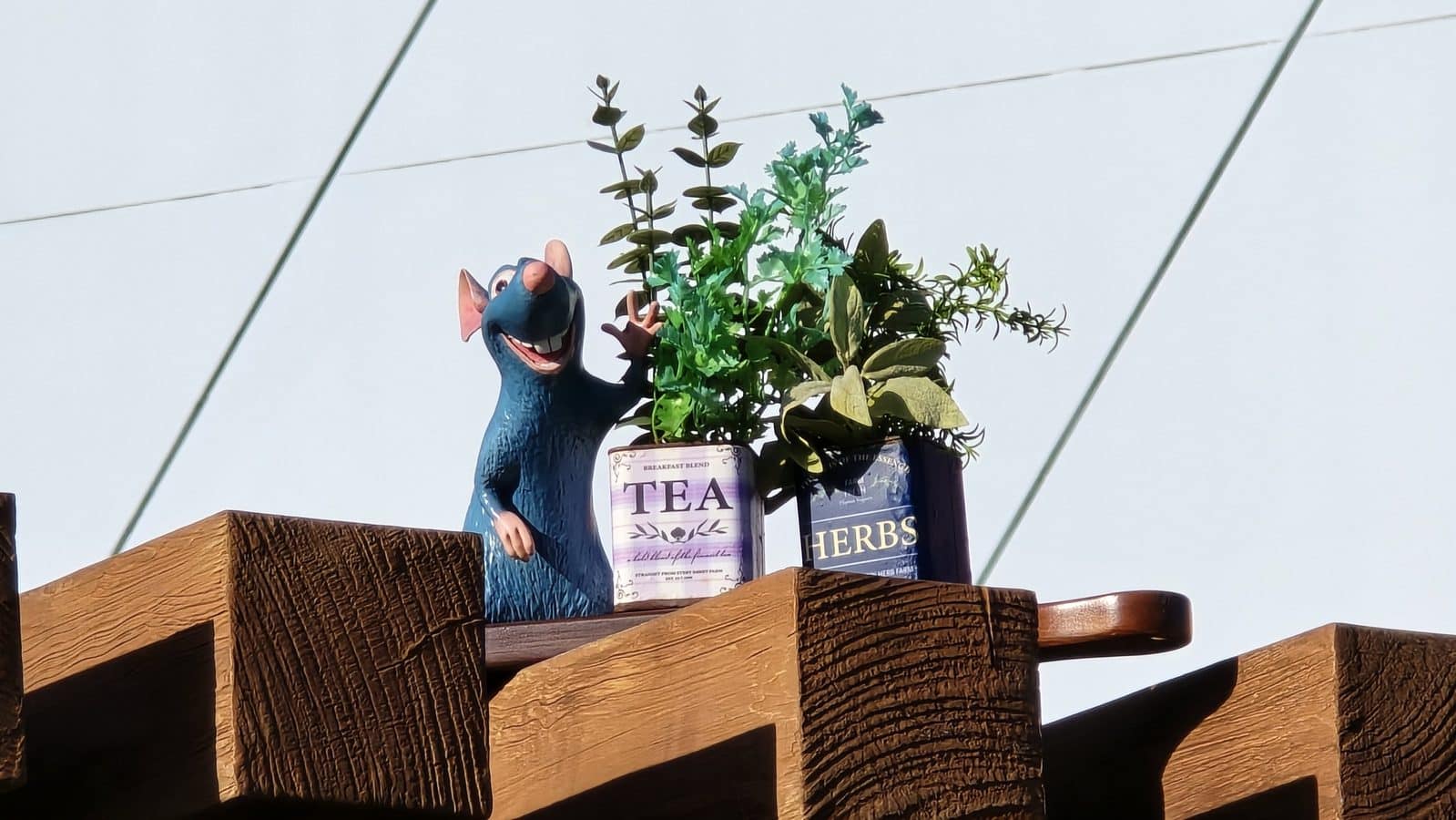 Blue cartoon rat statue beside potted herbs on a wooden ledge with tea and herbs containers visible and greenery spilling over the pots
