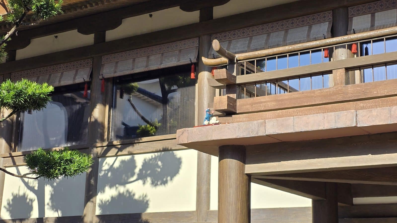 Wooden balcony with carved beams and decorative trim red tassels and a small statue on the railing Trees cast shadows nearby