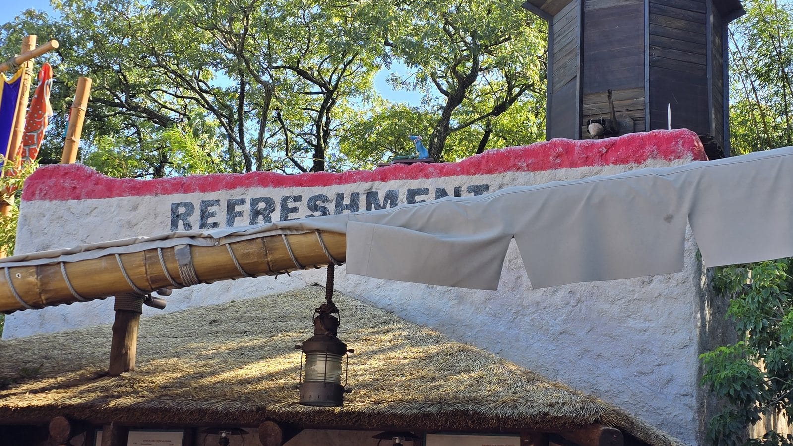 Outdoor refreshment stand with a white wall edged in pink thatched roof and bamboo posts amid green trees the word REFRESHMENT is visible on the wall