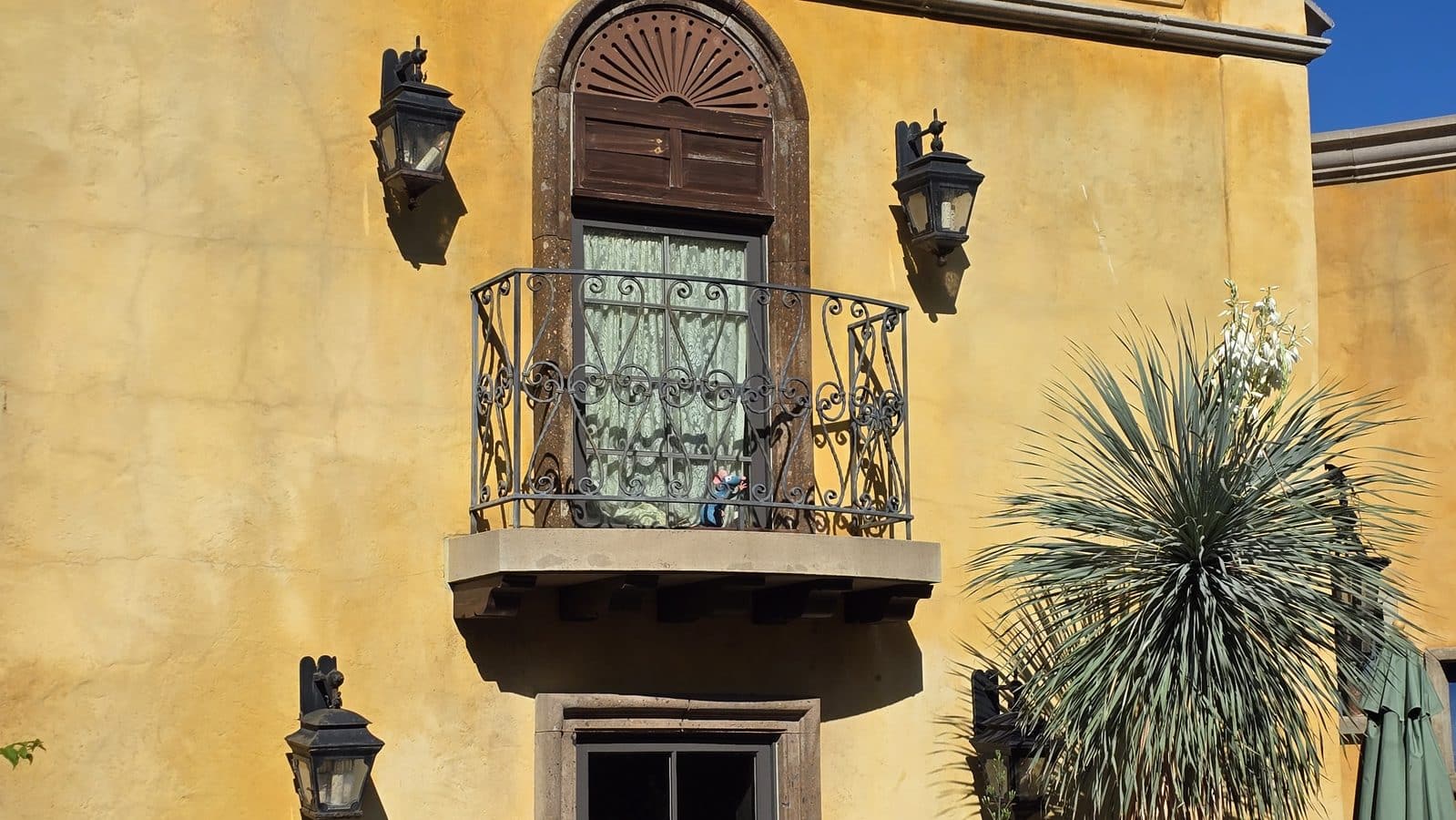 Yellow stucco building featuring a small decorative iron balcony with an arched window above and two wall lanterns on either side plus a palm plant to the right