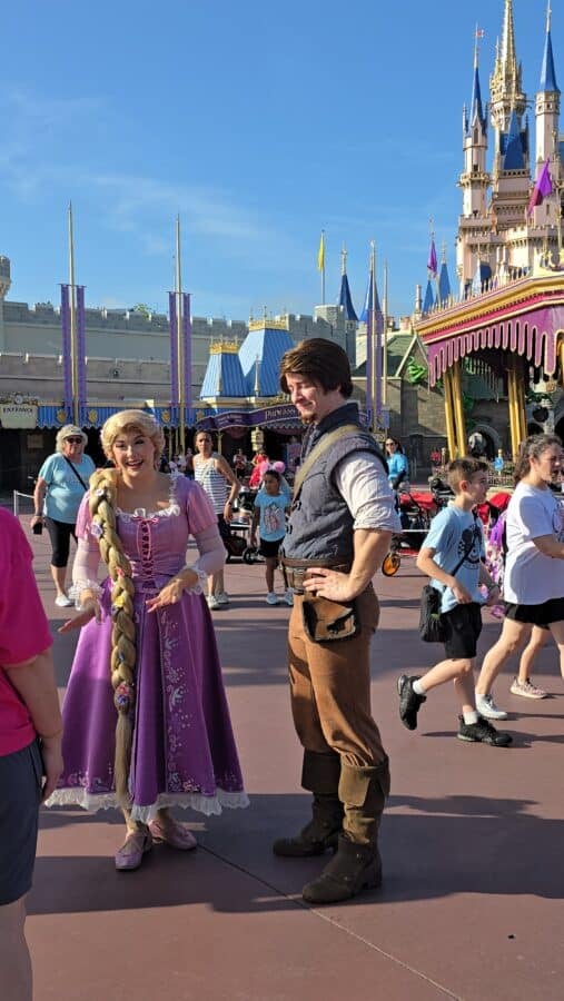 Two costumed characters pose for photos in a Disney park with a colorful castle in the background and visitors around them