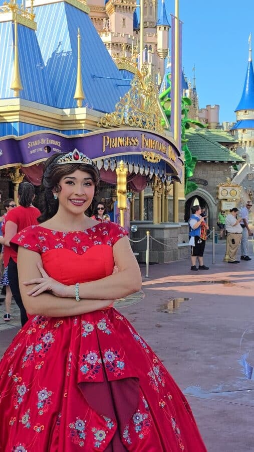 Smiling woman in a red floral ball gown and tiara poses in front of a castle entrance at a theme park