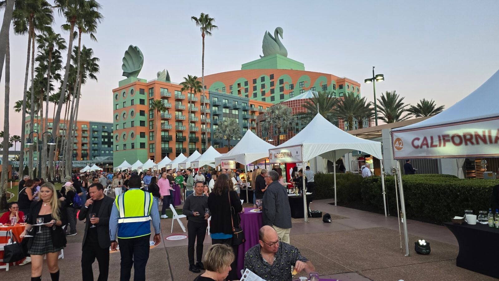 Crowd at an outdoor festival with white tents and a colorful hotel building in the background California banner on right palm trees framing the scene