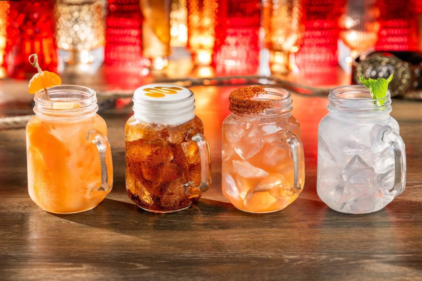 Four mason jar cocktails on a wooden bar each with ice orange juice drink with orange garnish dark iced beverage with whipped cream topping pink grapefruit drink with a chili salt rim and a clear mint garnished drink on the right