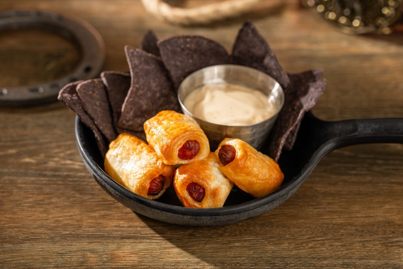 Sausage filled pastries with a creamy dip in a skillet surrounded by dark tortilla chips on a wooden table