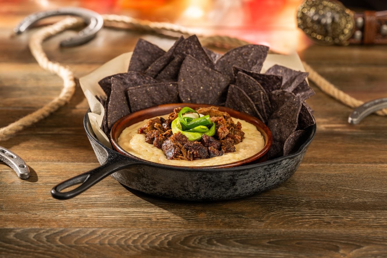 Cast iron skillet with creamy hummus topped by spiced beef and green onions surrounded by blue corn tortilla chips