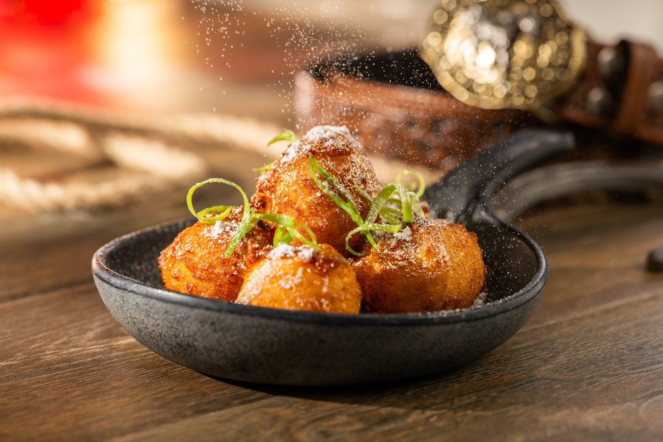Golden fried dough balls dusted with powdered sugar and garnished with thin green citrus zest in a dark bowl on a wooden table