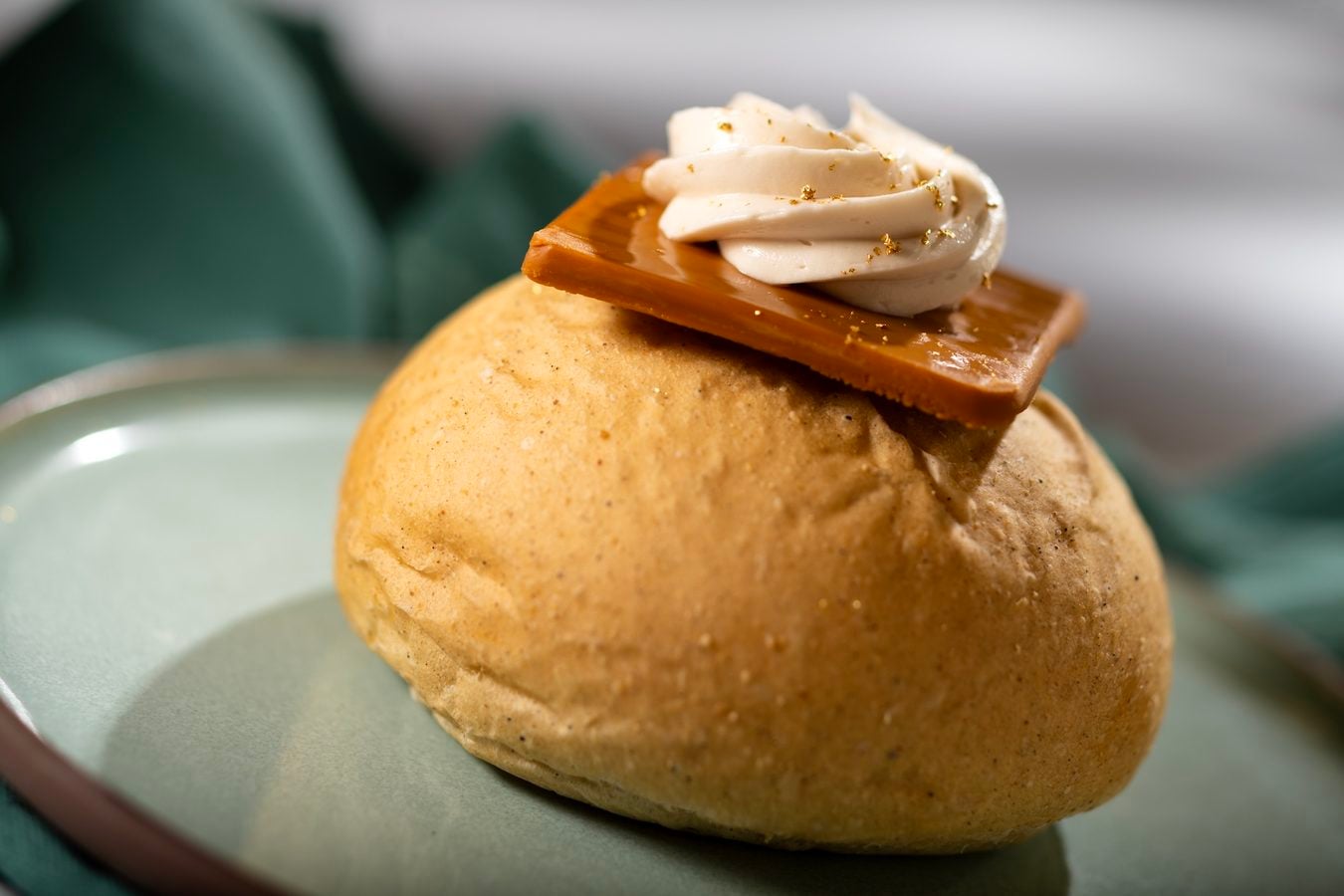 Round dessert bun topped with a glossy caramel shard and a swirl of whipped cream on a pale plate with gold flecks of garnish a close up shot edible dessert details included