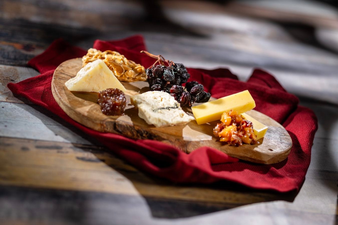 Wooden cheese board with assorted cheeses dried fruit grapes and a slice of yellow cheese on a red cloth