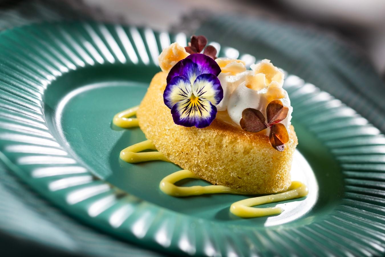 Slice of yellow cake topped with whipped cream candied fruit and an edible purple pansy on a green plate