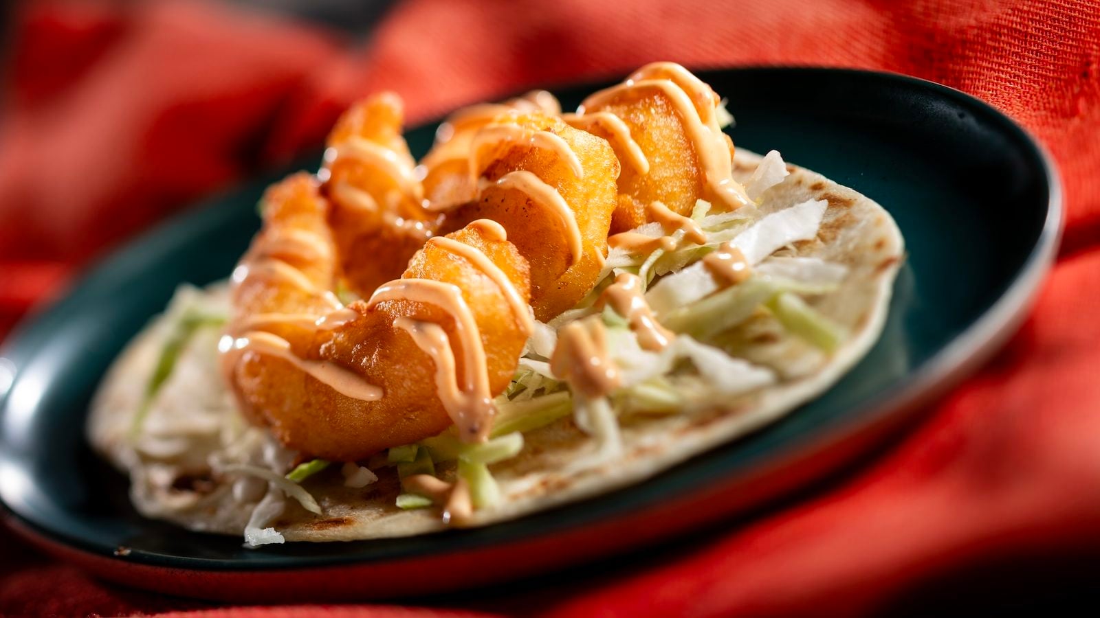 Taco on a soft tortilla filled with fried chicken shredded lettuce and drizzled sauce on a dark plate with a red tablecloth backdrop
