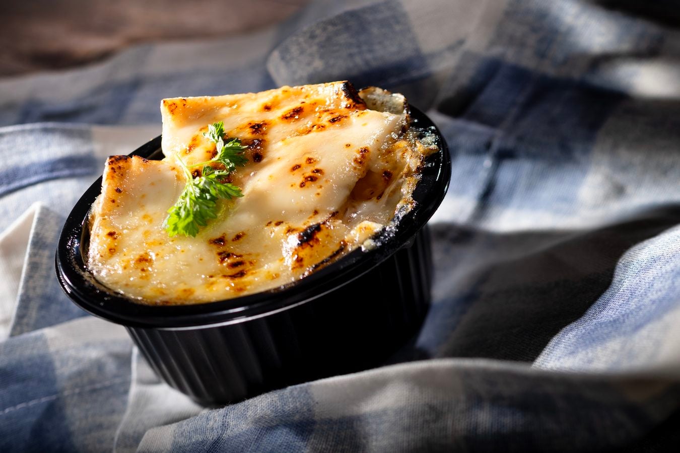 Melted cheesy gratin in a black ramekin garnished with a parsley sprig set on a blue cloth