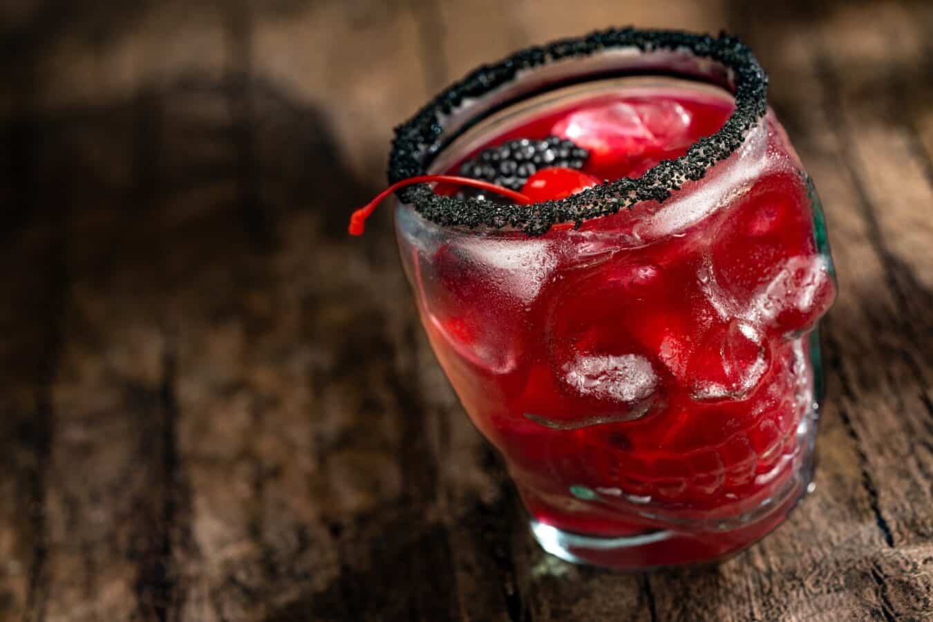 Red cocktail in a glass with a black sugar rim garnished with a red cherry and berries on a wooden table