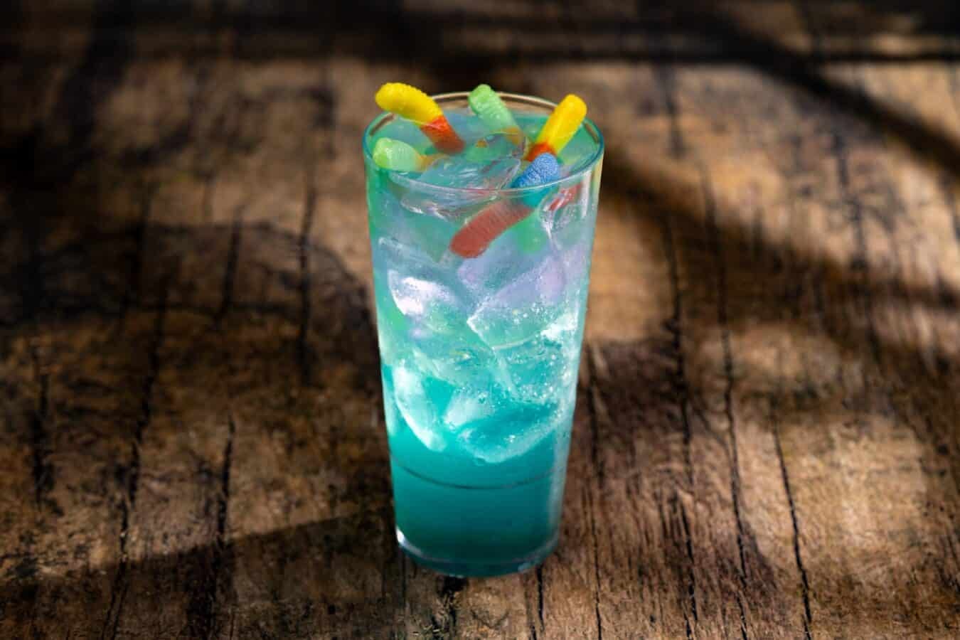 Tall blue cocktail in a highball glass with ice and colorful gummy worms on top set on a rustic wooden table