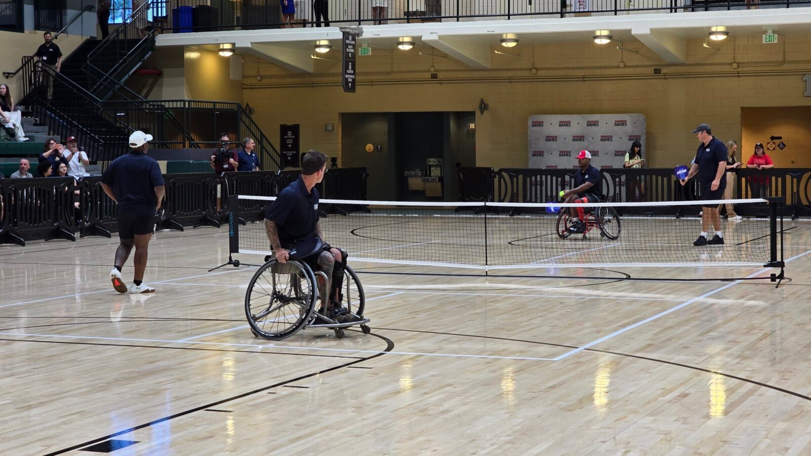 Wheelchair tennis players compete across a short indoor court with a net spectators are seated and watching from the sides