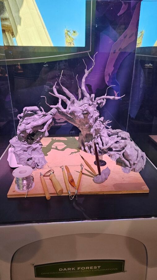 White gnarled driftwood sculpture with twisting branches on a wooden base displayed in a glass case with carving tools in the foreground