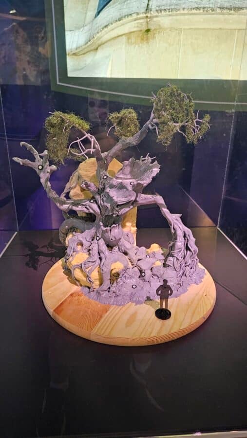 A sculpted bonsai like tree with twisted gray roots and branches moss tufts on the upper limbs displayed on a round wooden base in a glass case