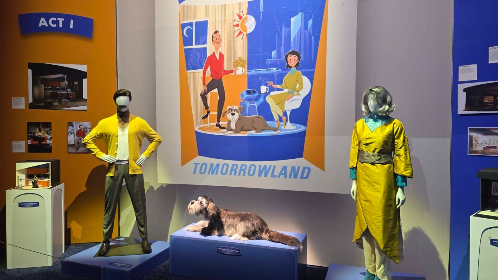 Museum display with two mannequins in bright yellow outfits beside a large Tomorrowland poster featuring a cartoon couple and a dog on a blue pedestal