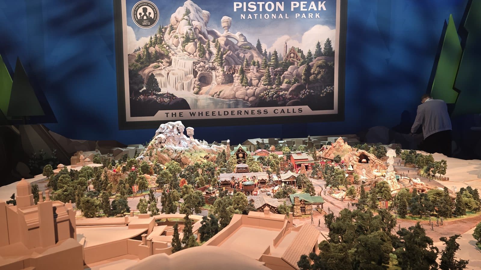 Detailed miniature mountain town diorama with forests rocks and buildings set in front of a large Piston Peak National Park poster