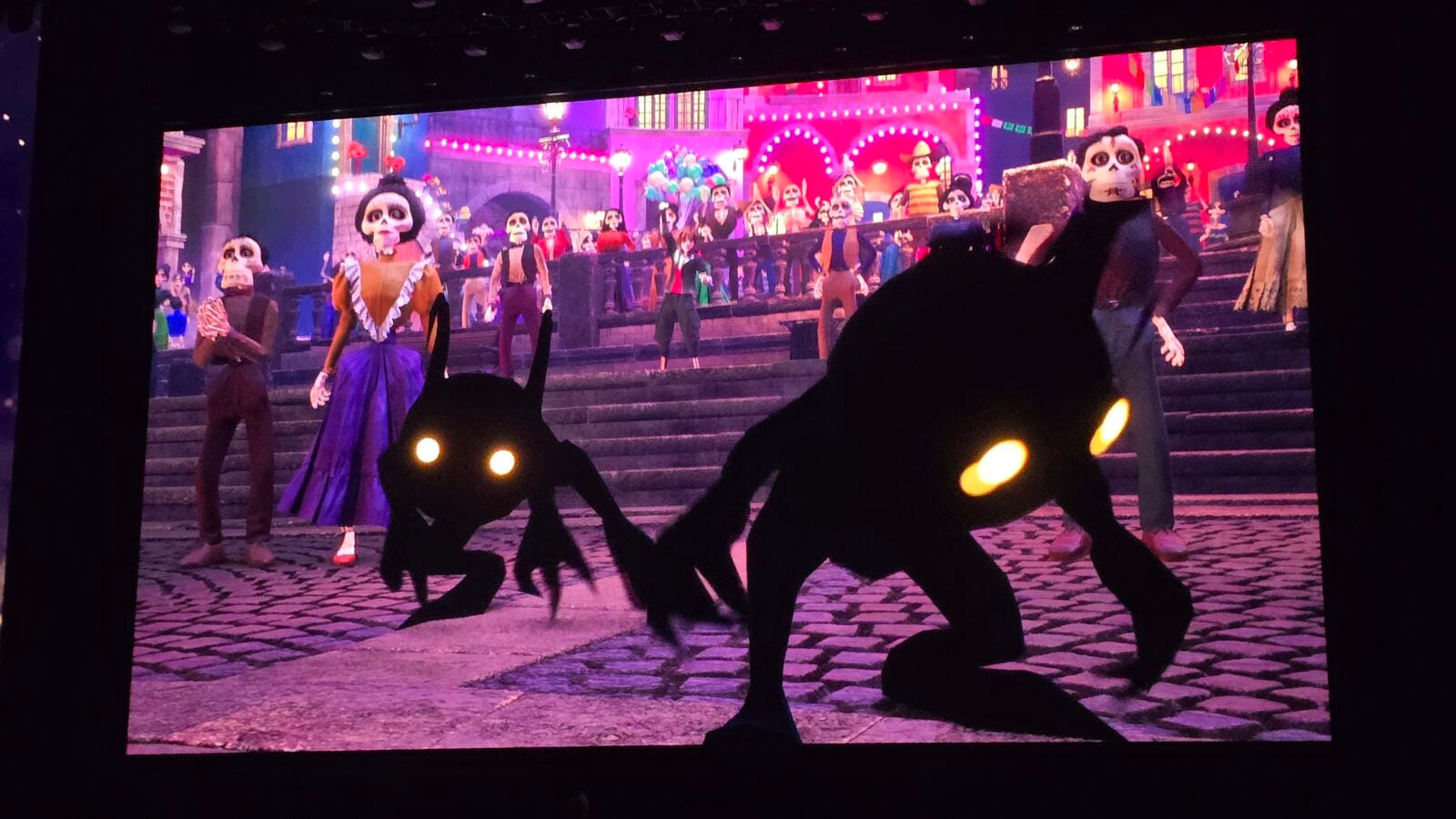 Colorful stage performance featuring skeleton dancers in bright outfits on steps with dark foreground silhouettes and glowing yellow eyes framing the scene