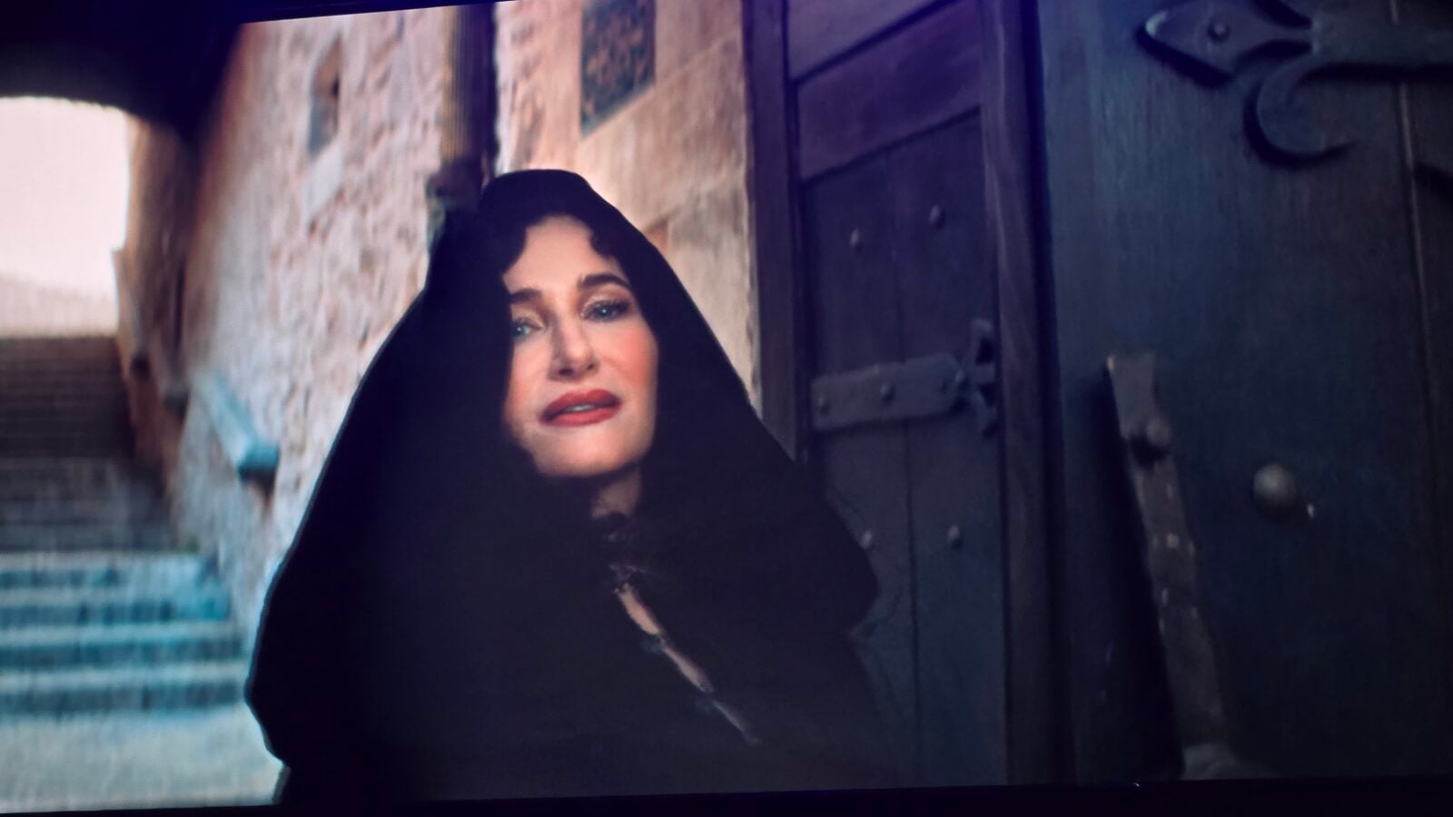 Woman in a black hooded cloak stands near a dark wooden door in a stone walled passage