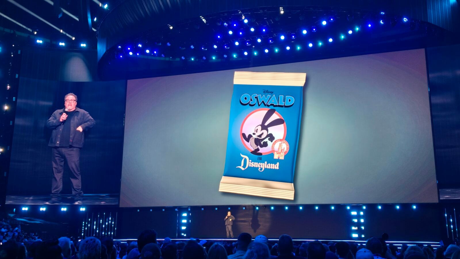Stage presentation with a man holding a microphone on the left and a large screen showing a Disney Oswald banner blue lighting and an audience in the foreground