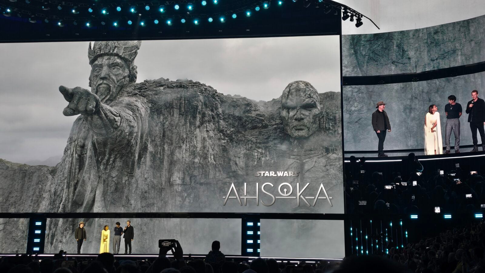 Stage audience watching a Star Wars Ahsoka presentation with a giant stone statue image on the screen and a panel of speakers on the right