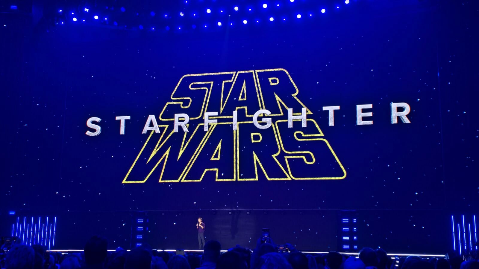 Stage presentation with a large Star Wars logo projected in yellow on a blue backdrop blue lights and an audience silhouette in foreground