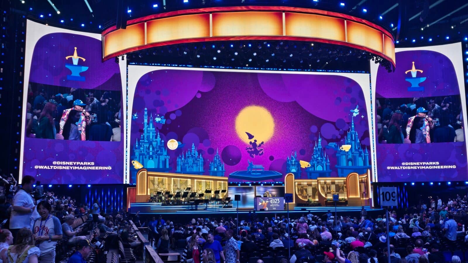 Large Disney themed stage with purple screens bright blue lights and an orchestra pit audience seated and watching a performance