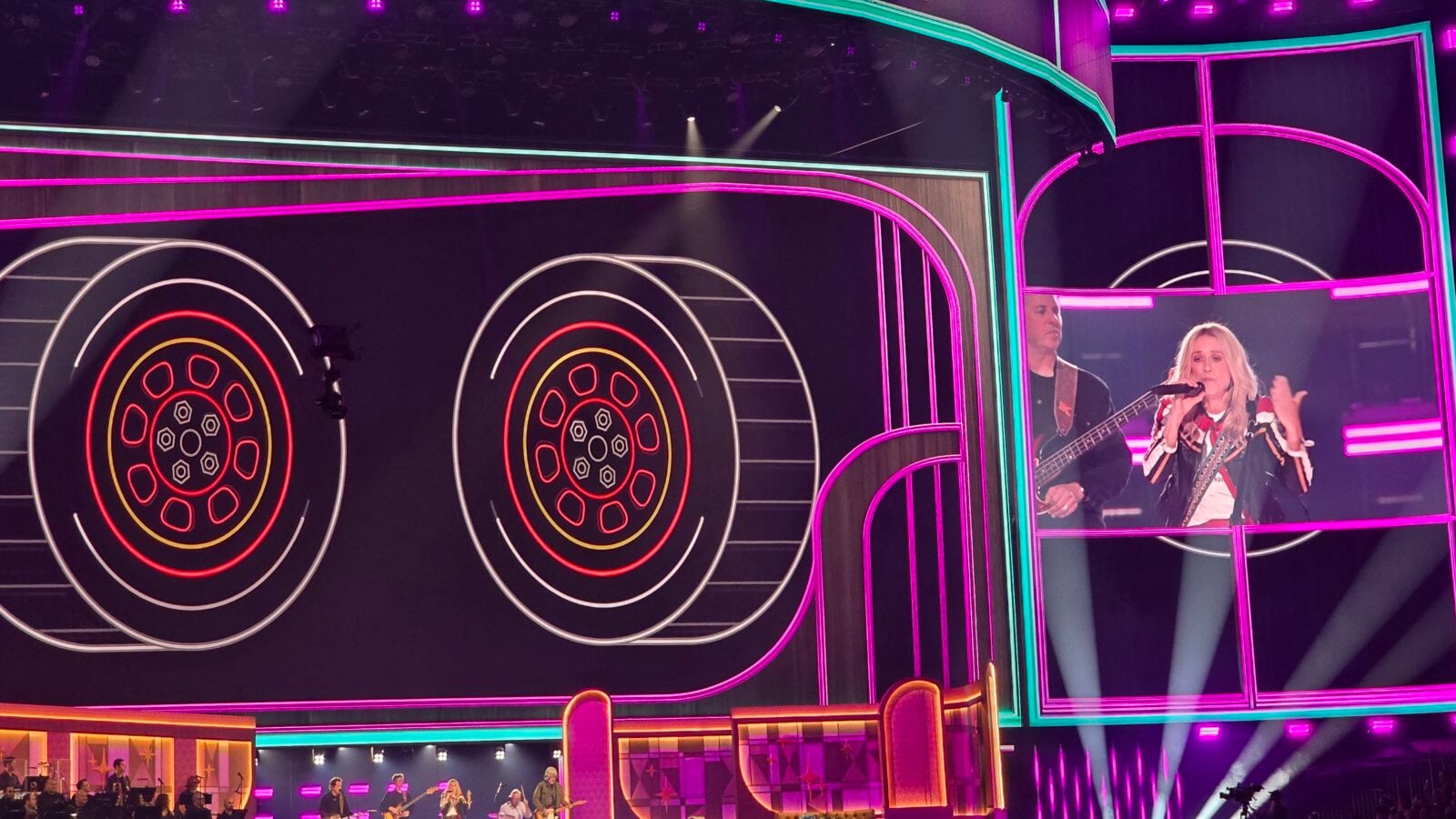 Concert stage with neon pink and purple lighting giant concentric circle wheel designs and a female singer at microphone with a guitarist in the background