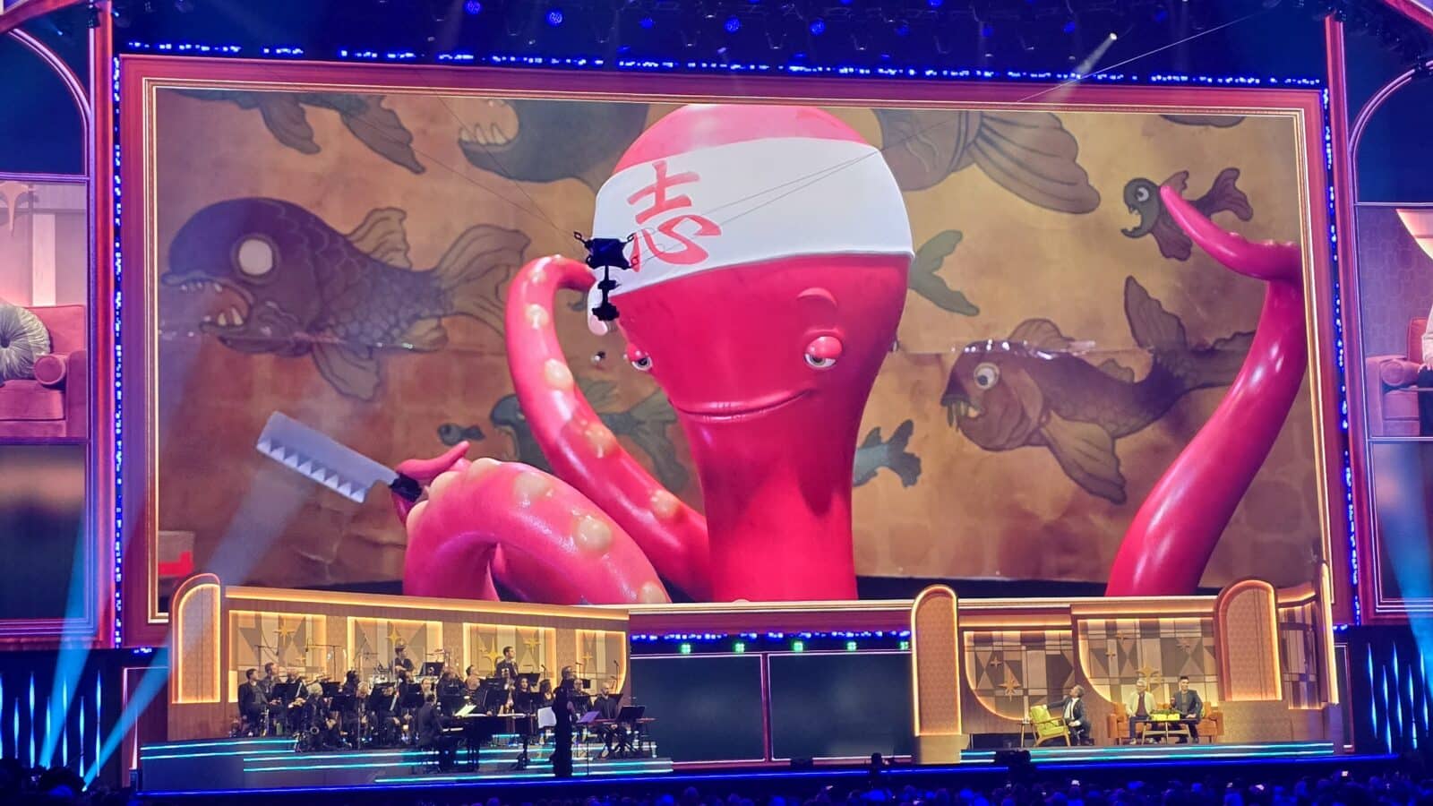 Giant pink octopus stage prop wearing a white headband with red characters holding a knife on a theater set with an orchestra below