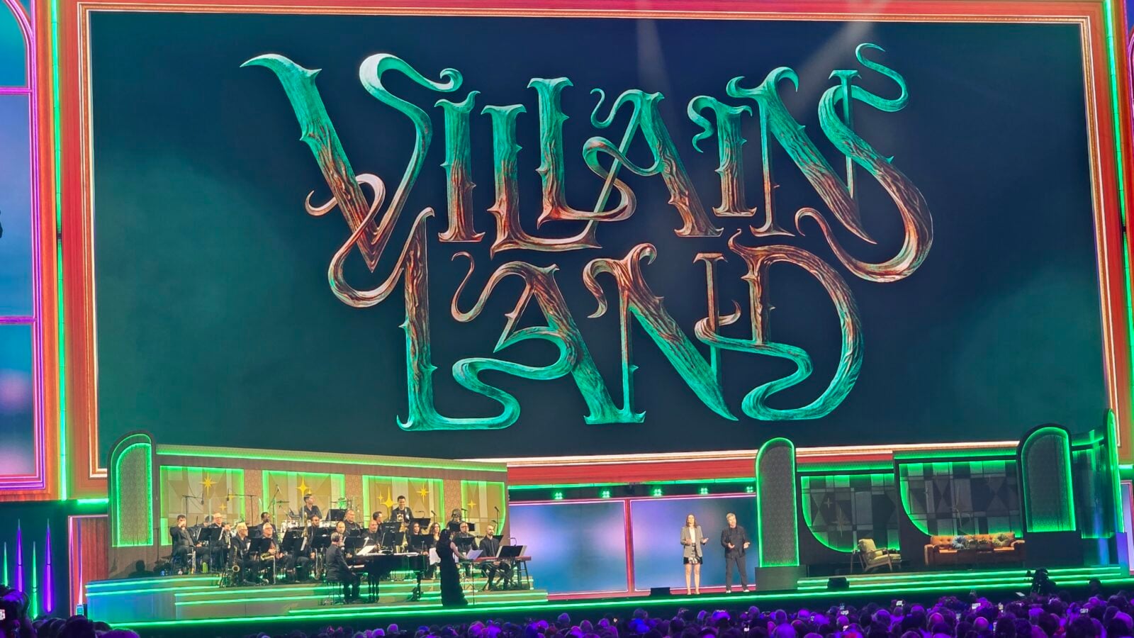 Theater stage for Wicked the Musical with a large stylized sign overhead an orchestra performing and two hosts at the front