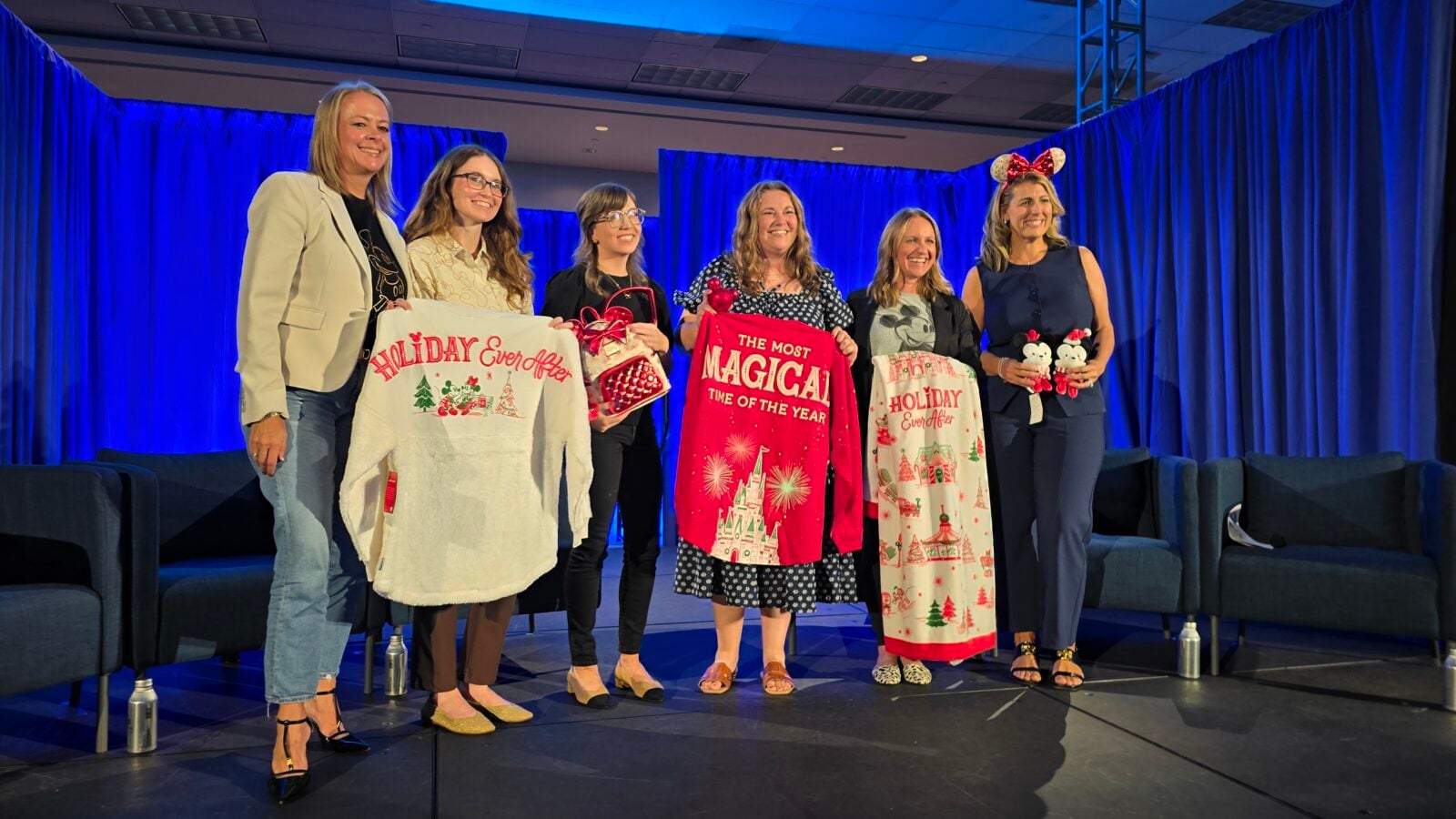Six women stand on a stage with blue curtains smiling holding festive holiday blankets and gifts