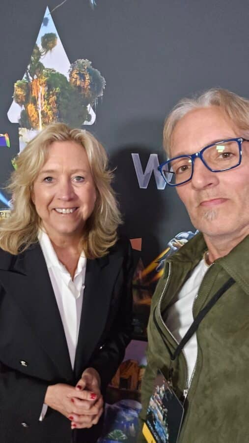 Smiling woman in a black blazer and white shirt posing with a man wearing blue glasses at an event backdrop for a selfie