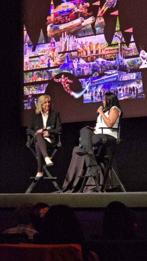Two women seated on stage in directors chairs each holding a microphone with a large colorful backdrop behind them