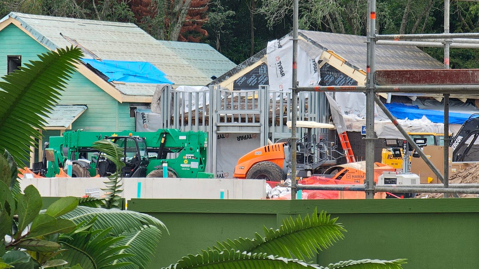 Construction site showing a partially built green house with scaffolding tarps and construction equipment in the foreground