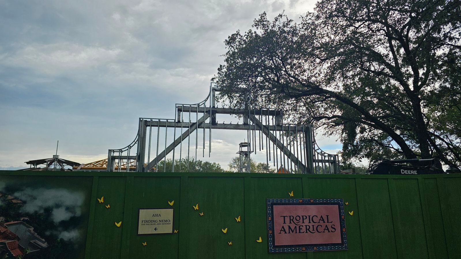 Construction site with exposed steel framing and a green fence a large tree on the right and a TROPICAL AMERICAS poster on the barrier