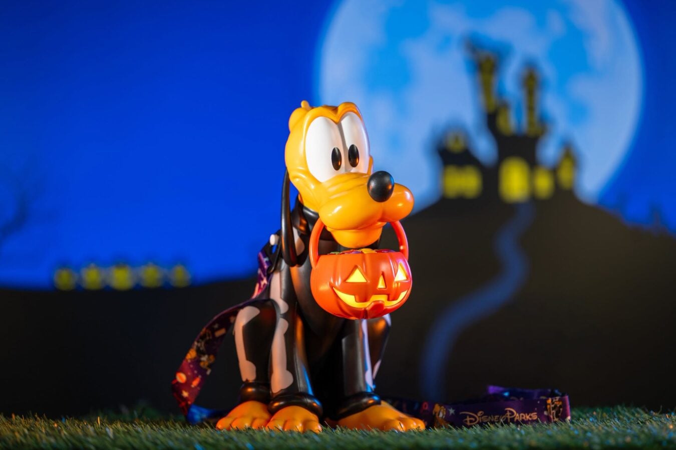 Costumed toy character with a carved pumpkin jack o lantern bucket standing on grass at night with a full moon and castle silhouettes in the background