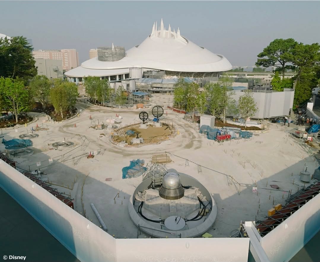 A construction site around a large white futuristic domed building with a crown like spire surrounded by trees and a circular plaza