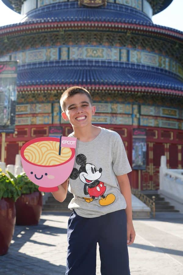 Smiling boy holding a pink ice cream cartoon cutout in front of a colorful temple facade with blue tiles and decorative panels behind him