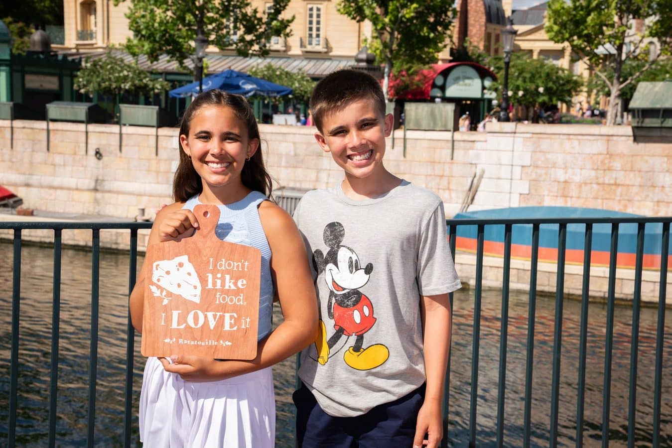 Two smiling kids stand by a railing along a riverfront one holding a wooden sign with text Mickey Mouse shirt on the other boy visible behind him