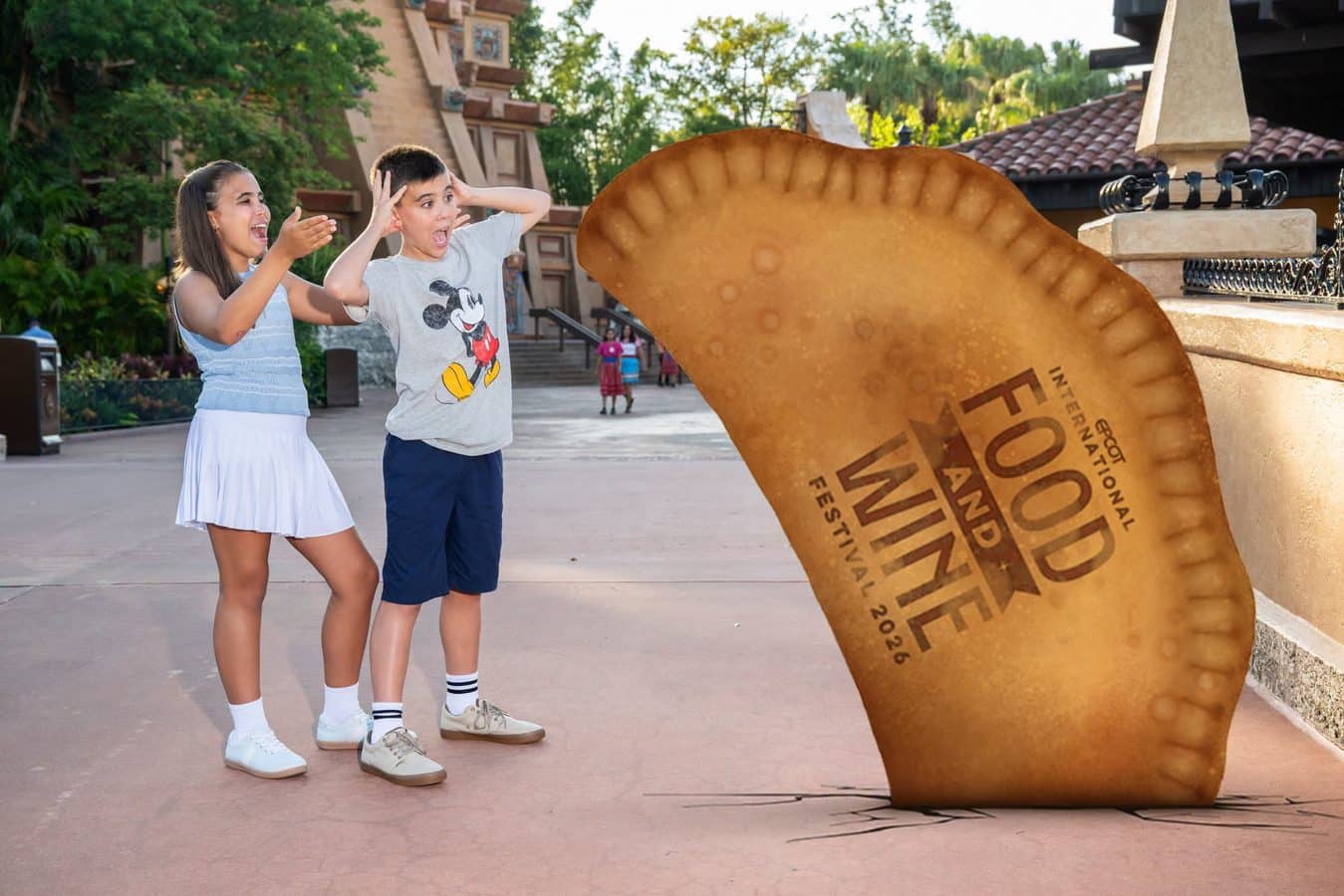 Two kids stand in a plaza laughing and gesturing as they react to a giant oversized gold coin sculpture nearby