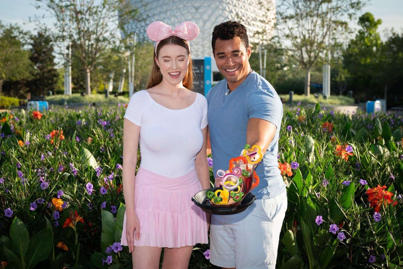 A smiling couple stands in a colorful flower garden woman in a white top and pink skirt with a pink bow headband man in a blue shirt holding a toy skateboard filled with colorful rings