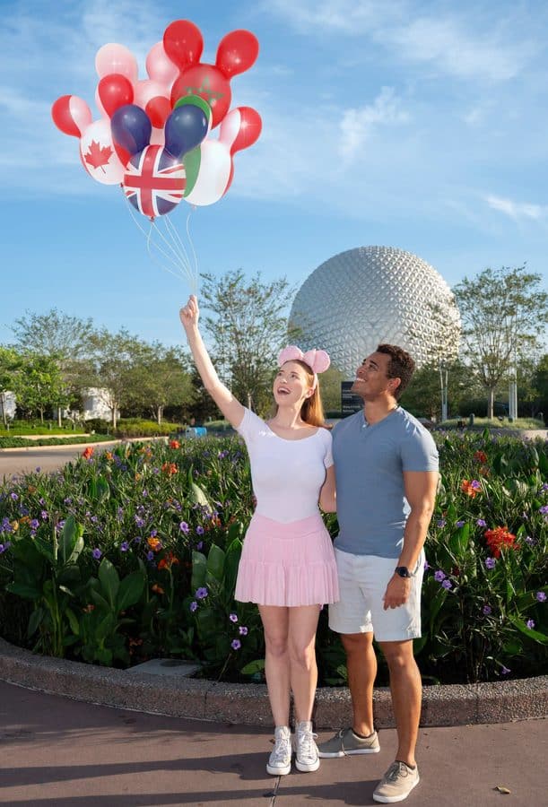 Couple in a park holds a cluster of international flag balloons as the woman points upward with a large geodesic sphere in the background