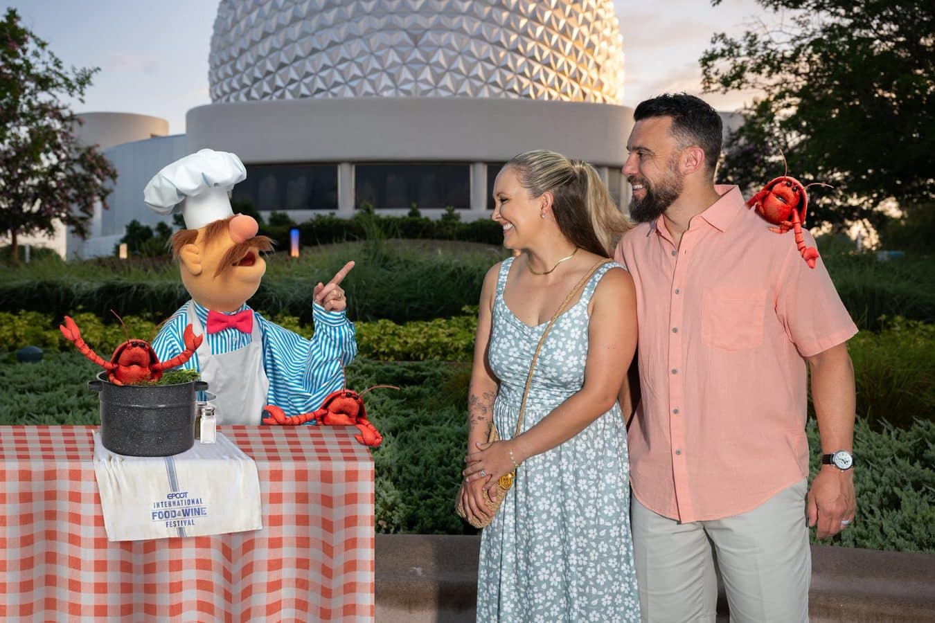 Couple chatting with a puppet chef and bright red lobsters on a checkered table at an outdoor festival