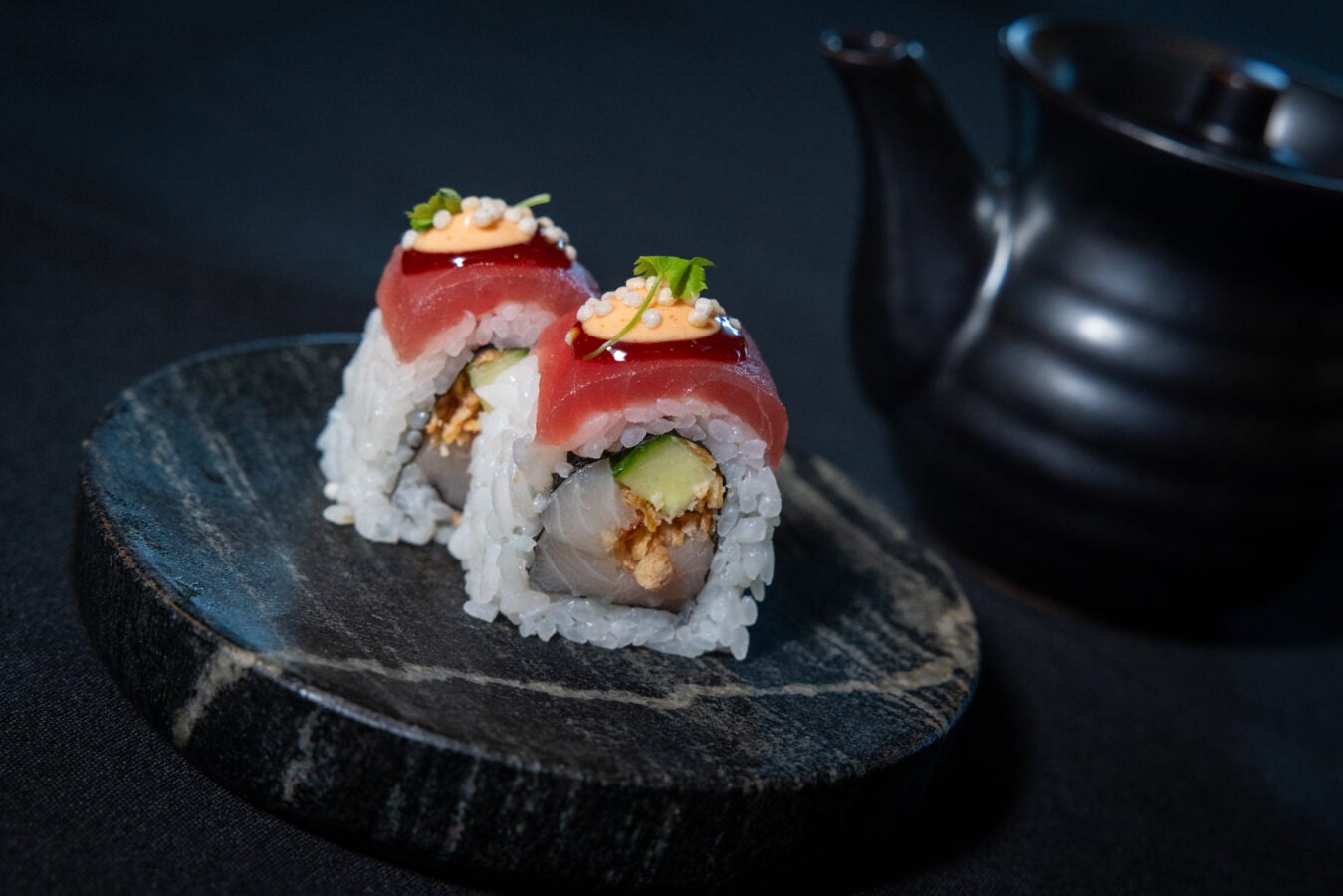 Two sushi rolls filled with avocado and crunchy filling topped with tuna slices orange sauce sesame seeds and a small green garnish on a dark plate