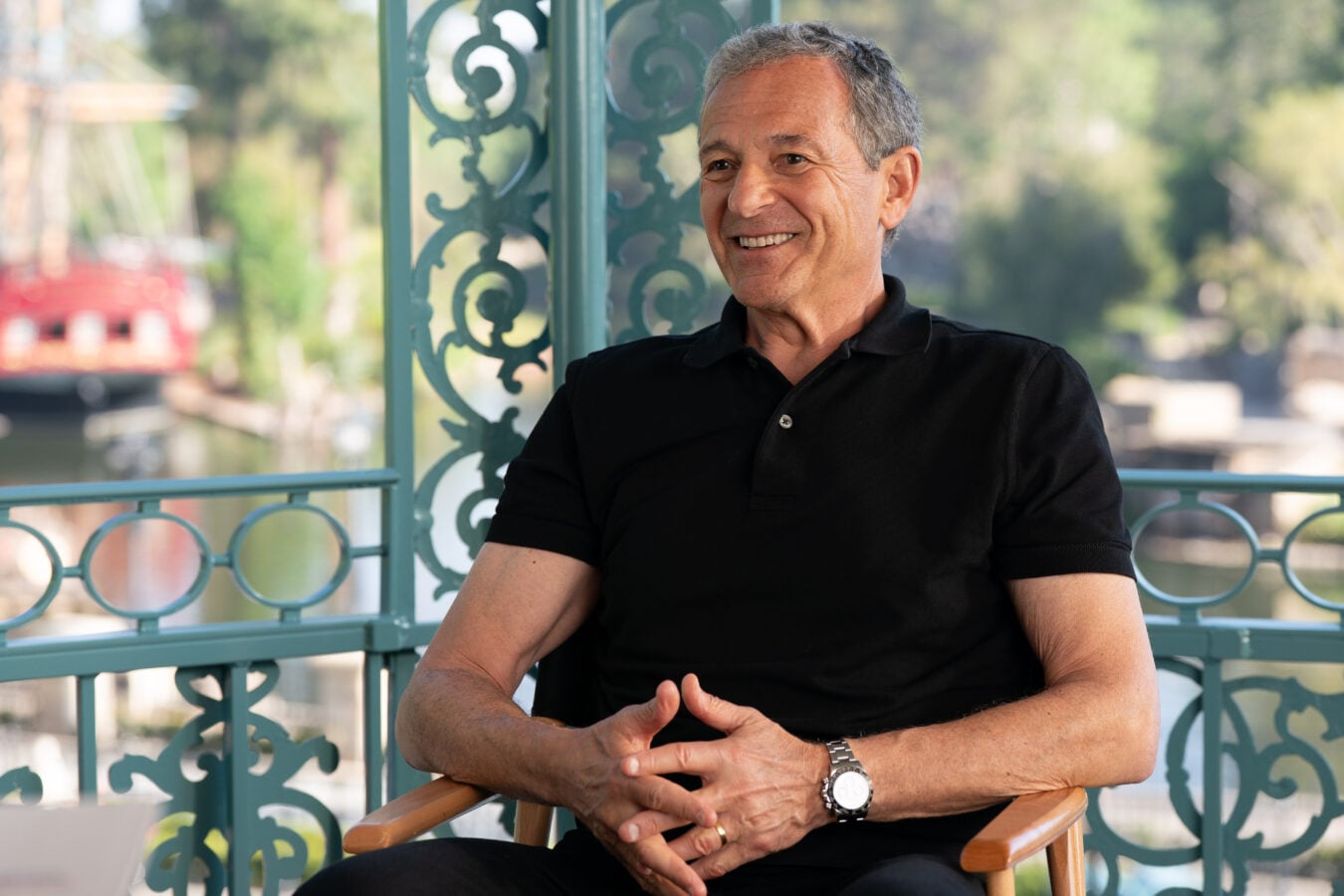 Smiling older man in a black polo seated outdoors beside teal decorative railing