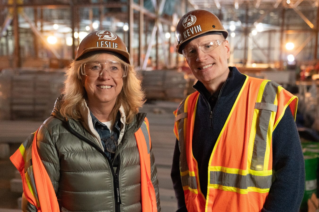 Two construction workers Leslie and Kevin smile at the camera in hard hats and reflective vests on a job site