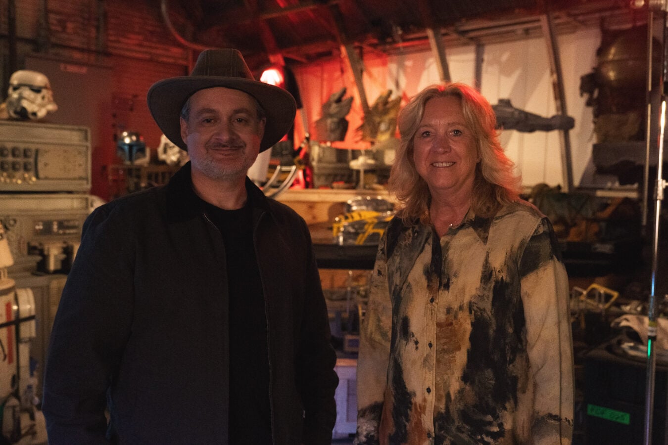 Two adults stand in a cluttered dimly lit workshop man wears a wide brimmed hat woman in a paint splattered shirt both smiling