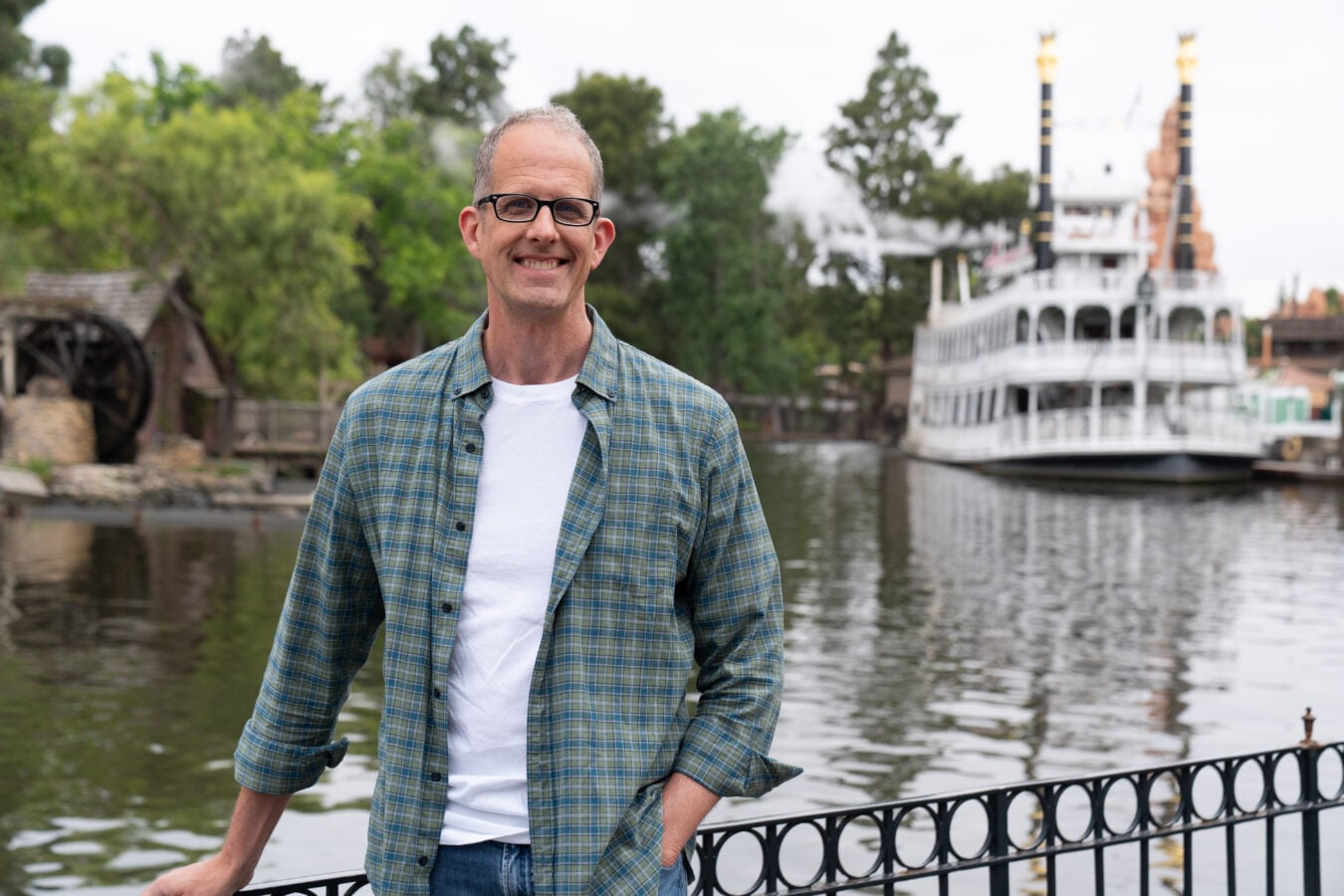 Smiling man in a green plaid shirt stands by a railing on the water with a white paddlewheel boat in the background